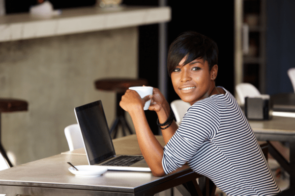 woman holding a cup of coffee, seated in front of laptop, looking at viewer